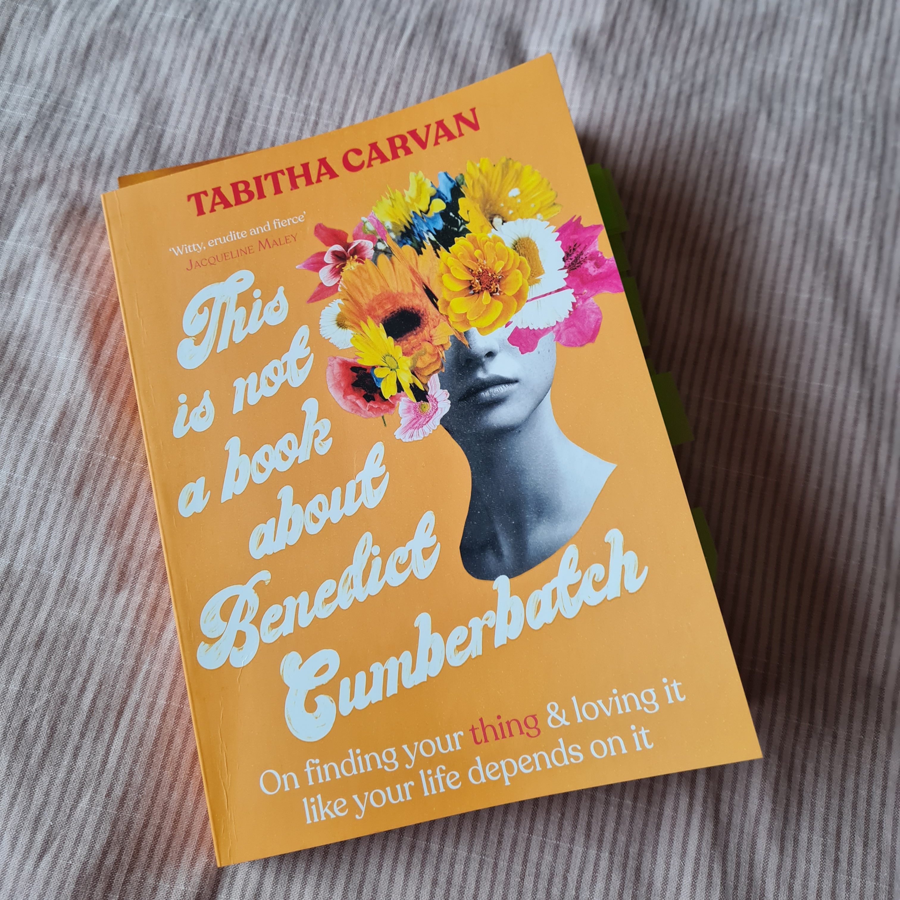 The book "This is not a Book about Benedict Cumberbatch" is sitting on a light pink blanket. The cover of the book is orange and has a black and white picture of a woman's face with flowers coming out of the top of her head.